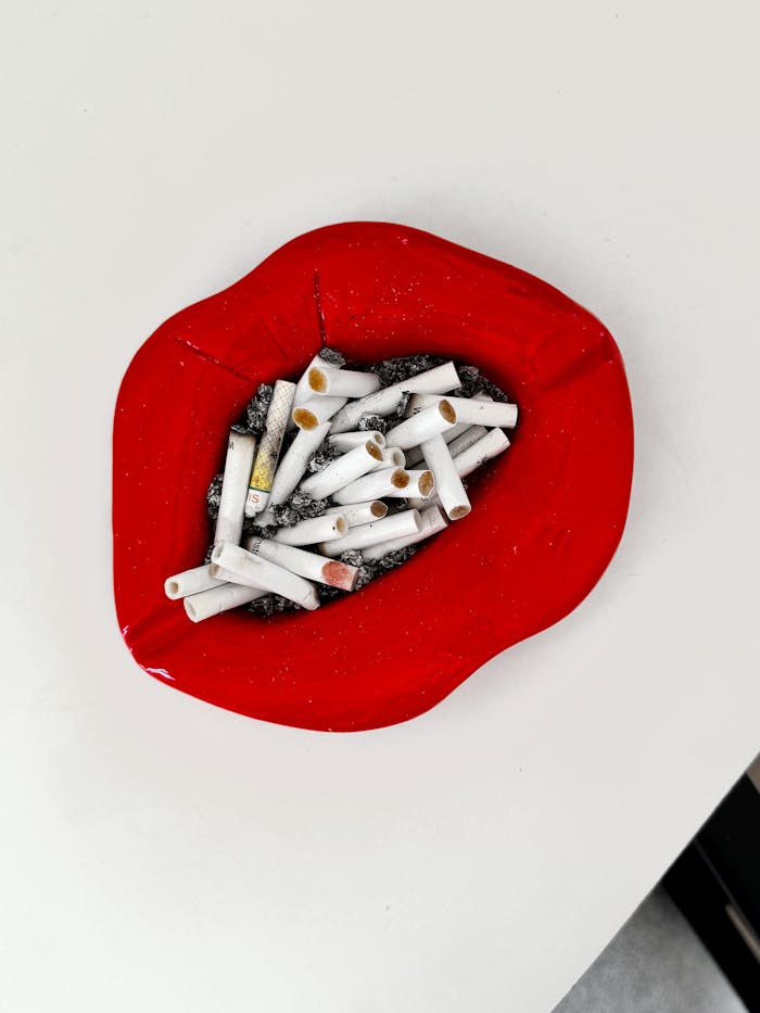 gallery-3 Close-up of a red lip-shaped ashtray filled with cigarette butts symbolizing smoking addiction and health risks.