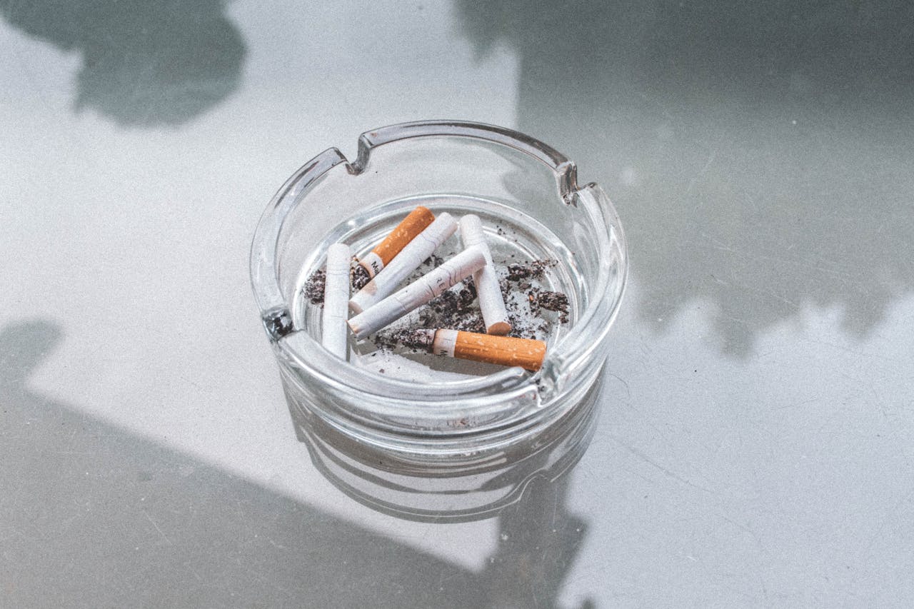 services-01 A close-up view of used cigarette butts in a glass ashtray on a reflective surface, highlighting indoor smoking.
