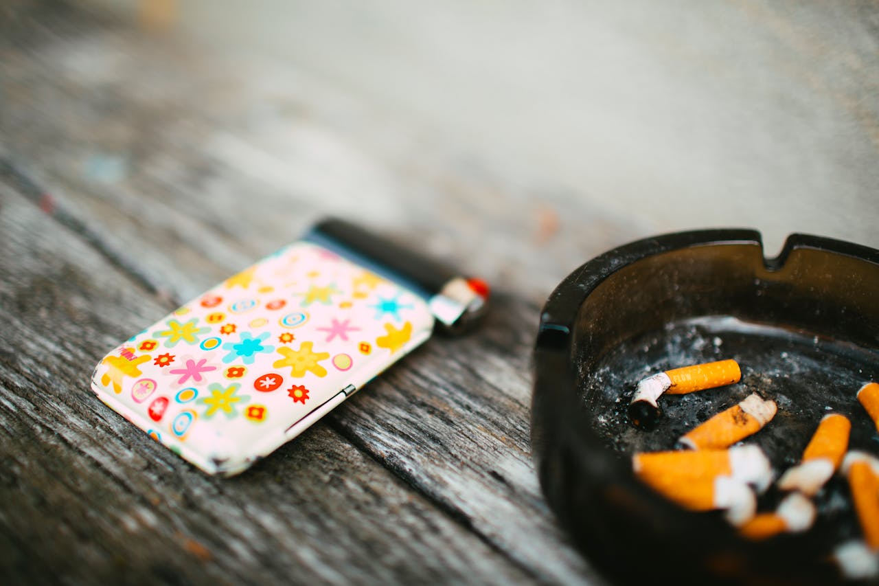 hero-img-02 A vibrant lighter sits next to a black ashtray filled with cigarette butts on a wooden surface.