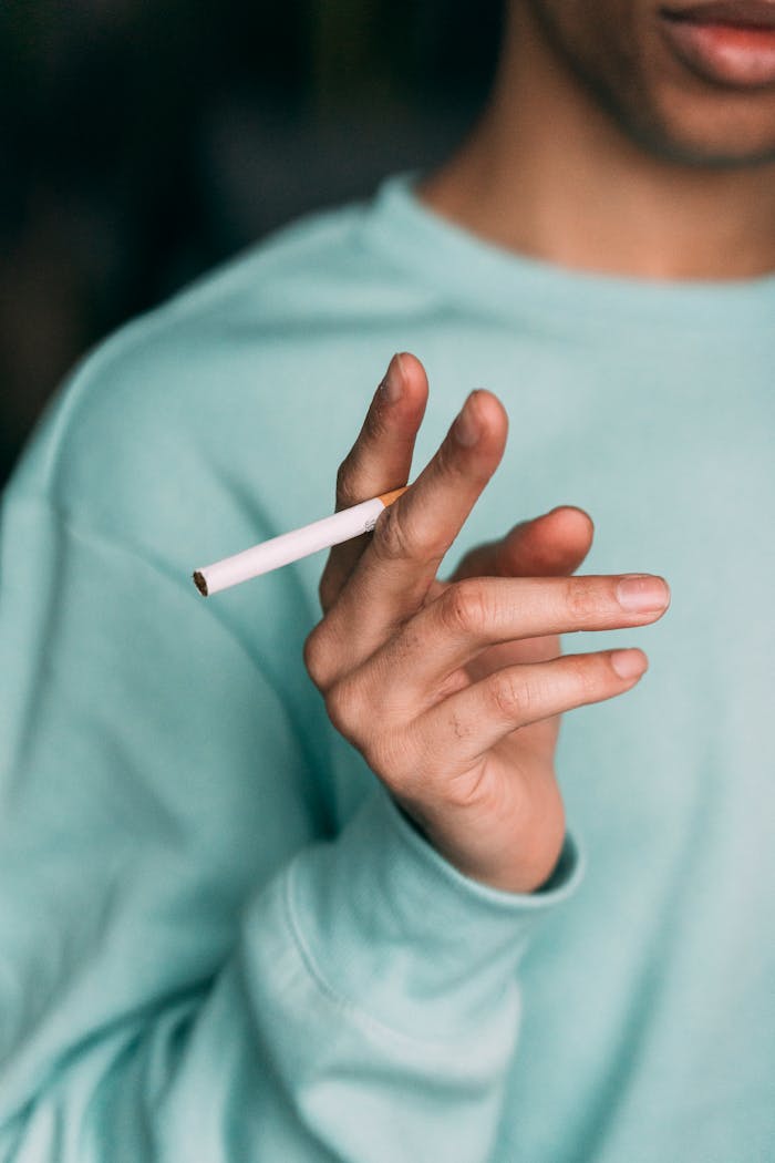 Crop anonymous male smoker in casual clothes with unlit cigarette in hand standing in light room at home during smoking